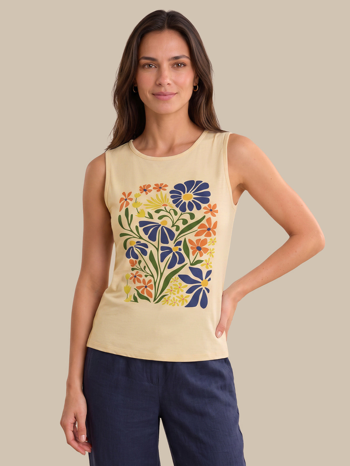 A woman modeling a sleeveless cream-colored tank top with a colorful floral print on the front, paired with navy blue pants, set against a solid beige background.