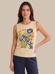 A woman modeling a sleeveless cream-colored tank top with a colorful floral print on the front, paired with navy blue pants, set against a solid beige background.