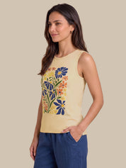 A woman modeling a sleeveless cream-colored tank top with a colorful floral graphic on the front, paired with navy blue pants, set against a solid beige background.