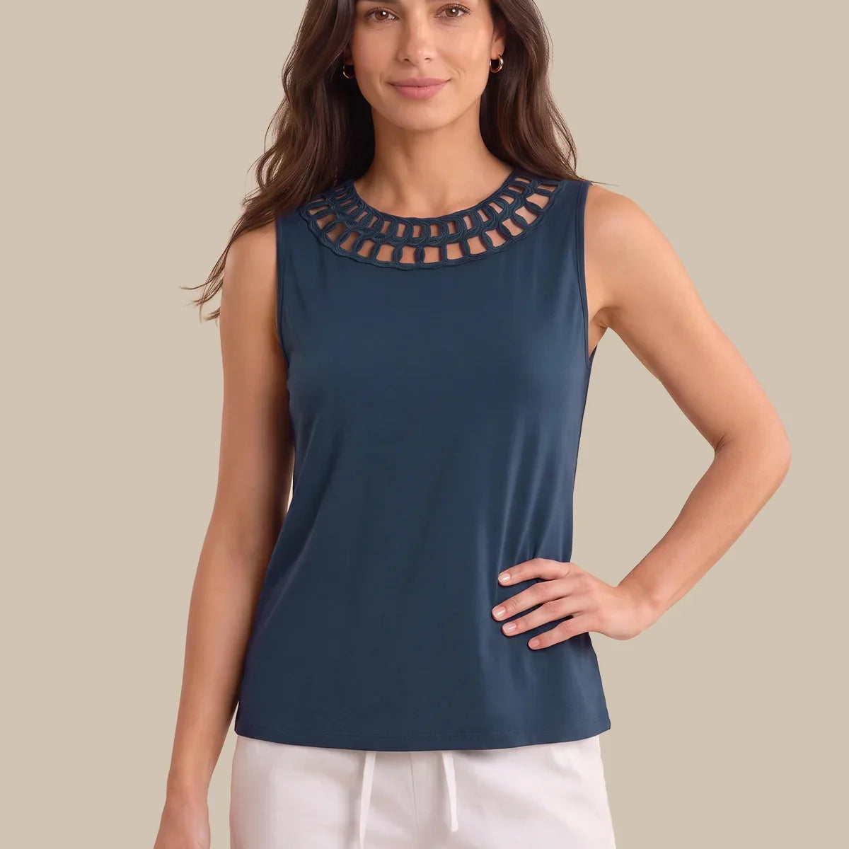 A woman modeling a sleeveless navy blue top with a decorative cutout neckline, paired with white drawstring pants, against a solid beige background.