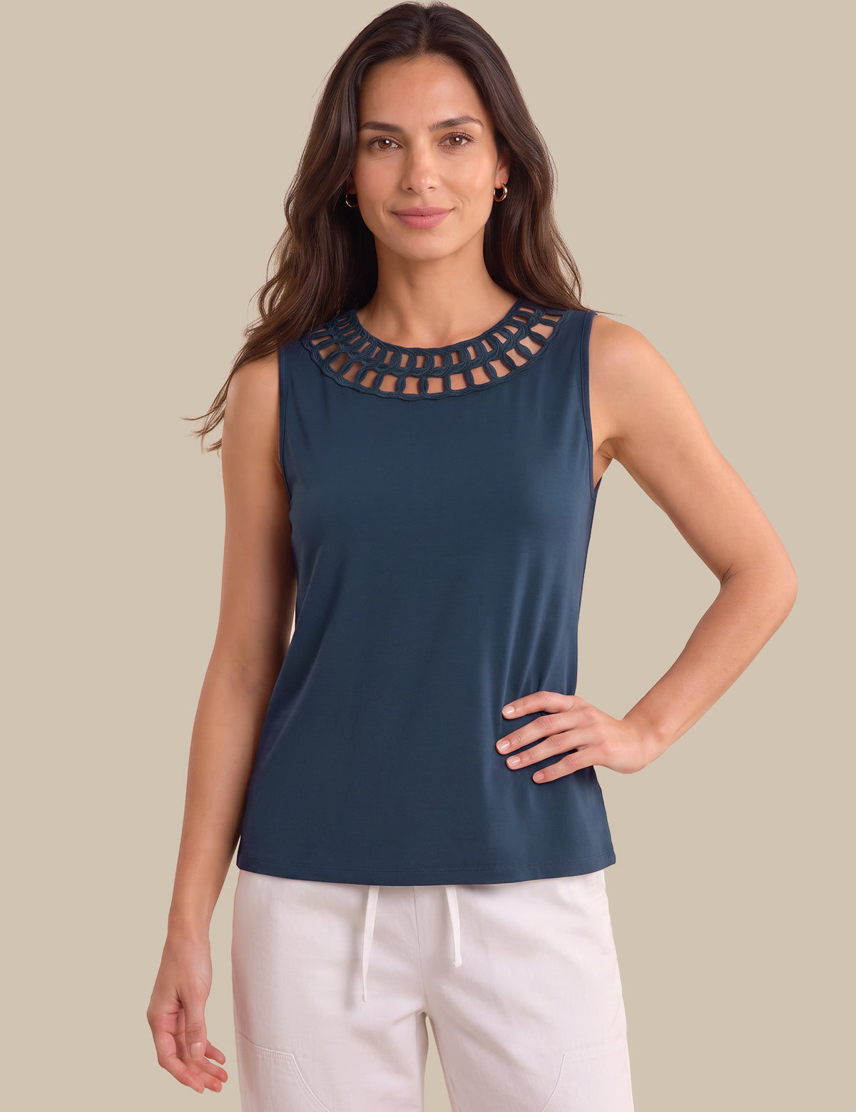 A woman modeling a sleeveless navy blue top with a decorative cutout neckline, paired with white drawstring pants, against a solid beige background.