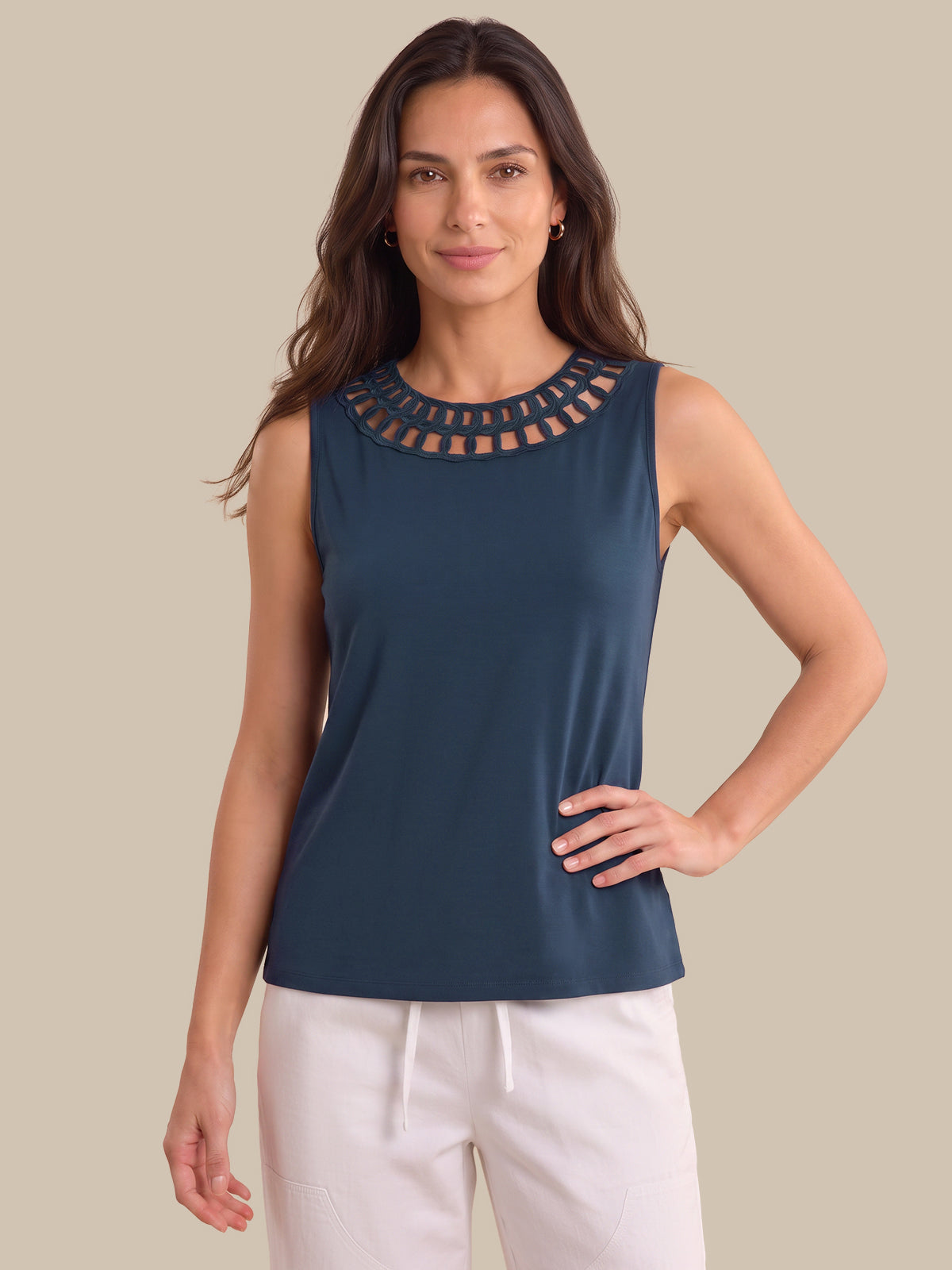 A woman modeling a sleeveless navy blue top with a decorative cutout neckline, paired with white drawstring pants, against a solid beige background.
