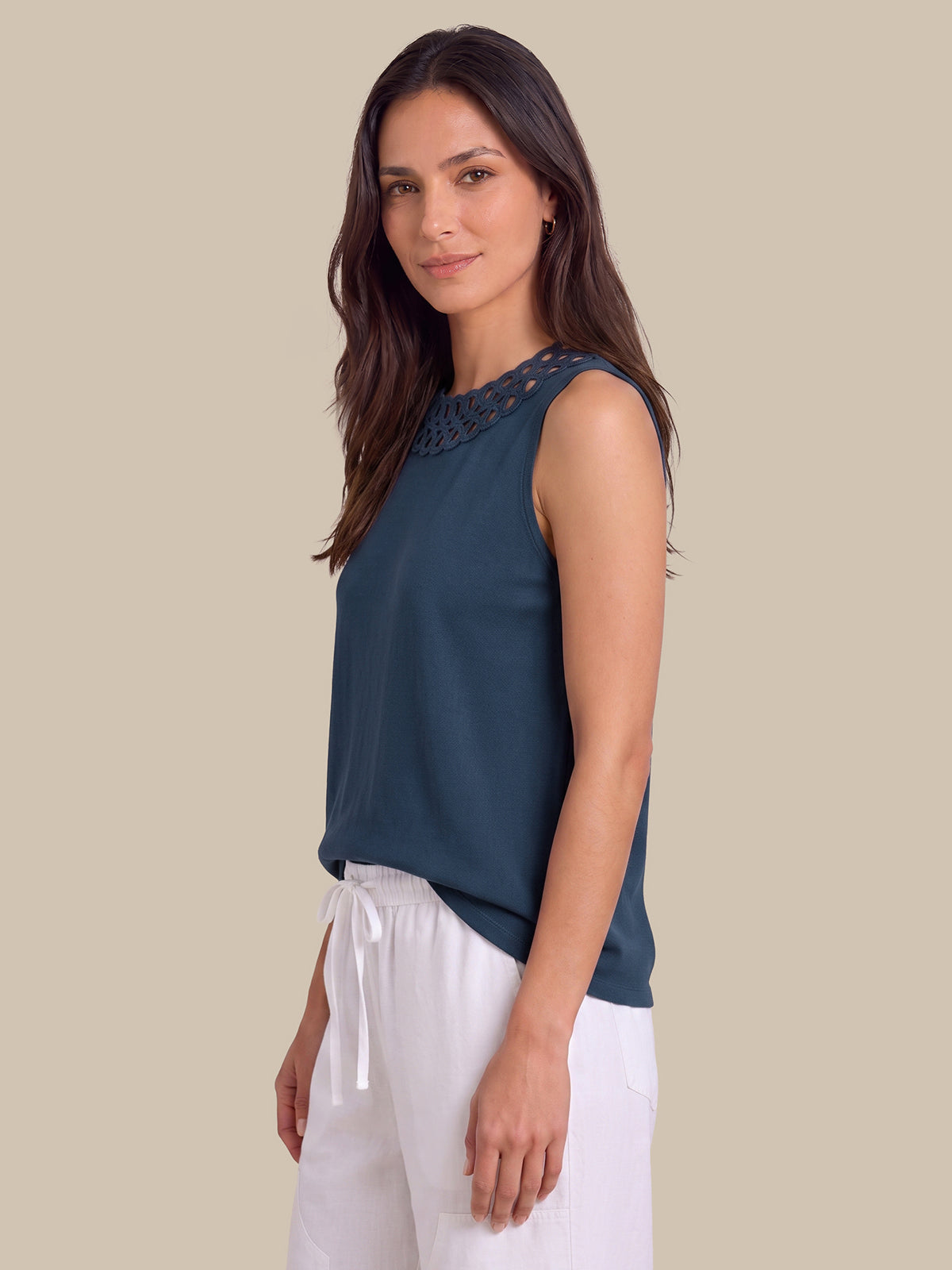 A woman modeling a sleeveless dark teal top with a lace-embroidered neckline, paired with white drawstring pants, against a solid beige background.