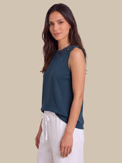 A woman modeling a sleeveless dark teal top with a lace-embroidered neckline, paired with white drawstring pants, against a solid beige background.