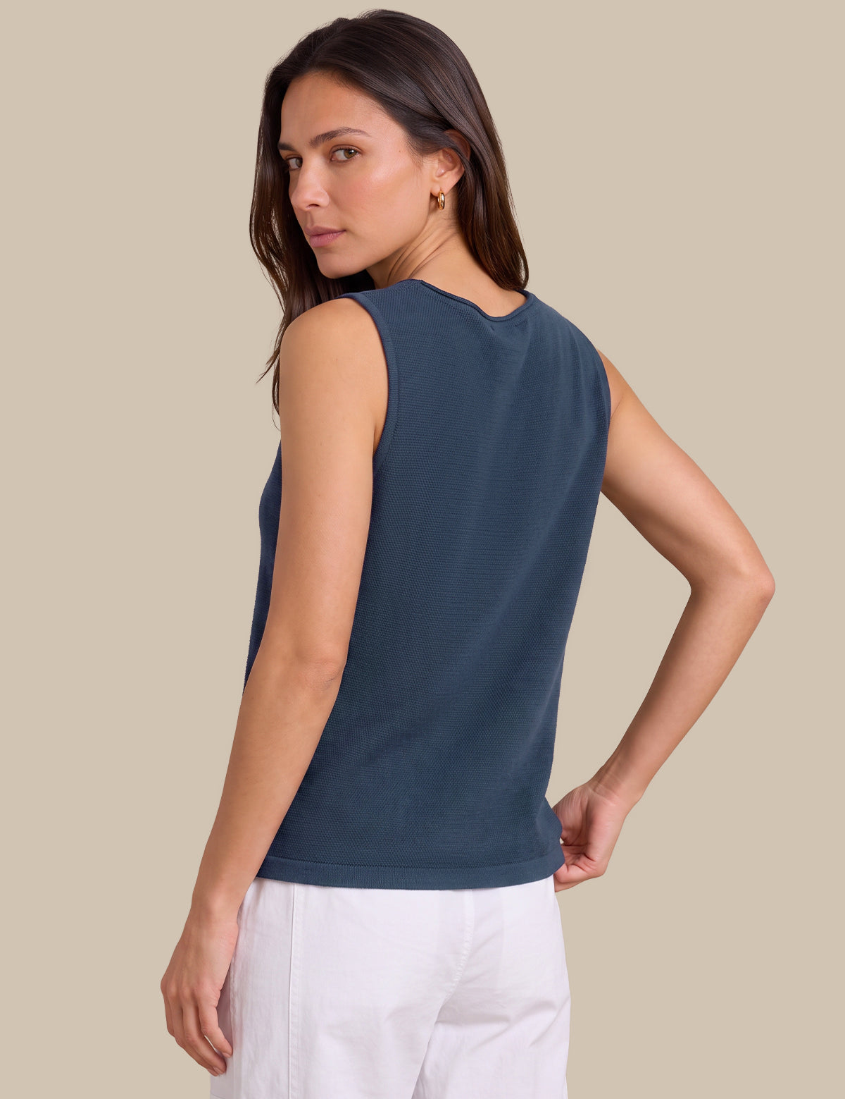 A woman wearing a sleeveless dark slate blue top and white pants, shown from a three-quarters back view against a solid beige background.