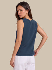 A woman wearing a sleeveless dark slate blue top and white pants, shown from a three-quarters back view against a solid beige background.