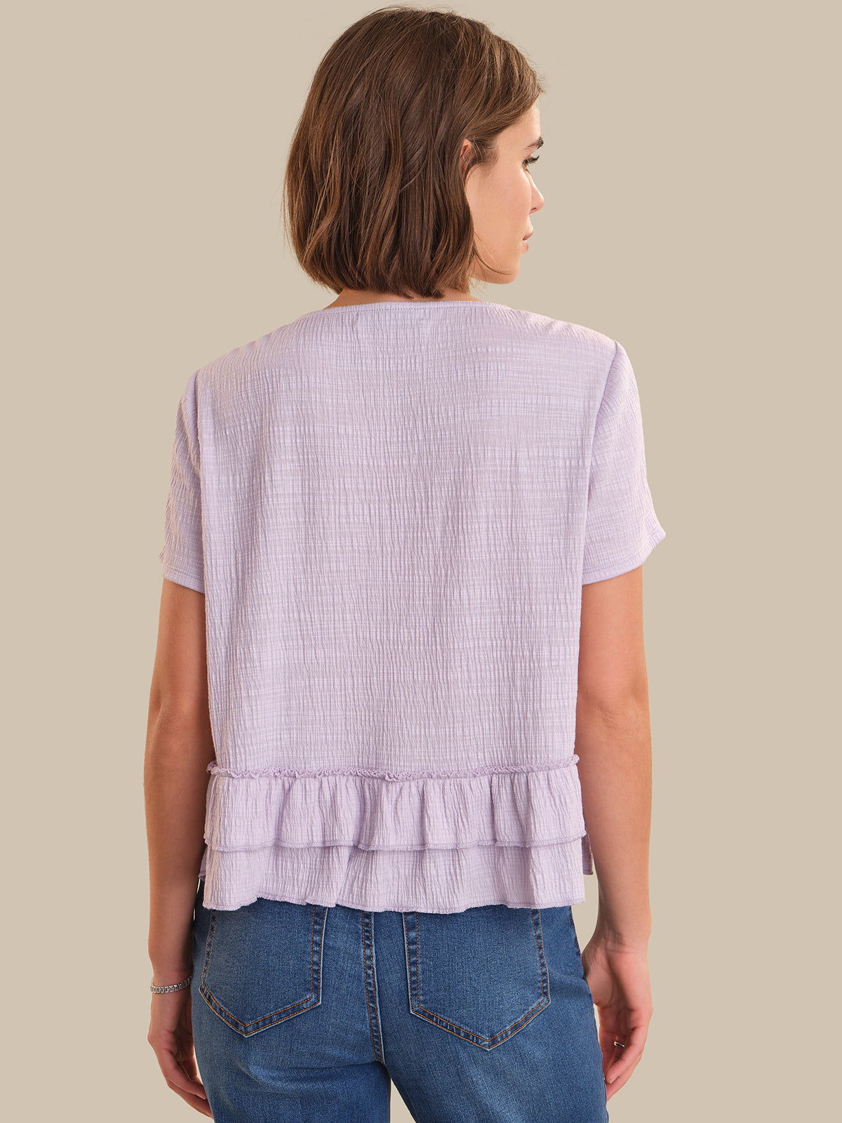 Woman wearing a light purple textured top with ruffled details and blue jeans on a beige background