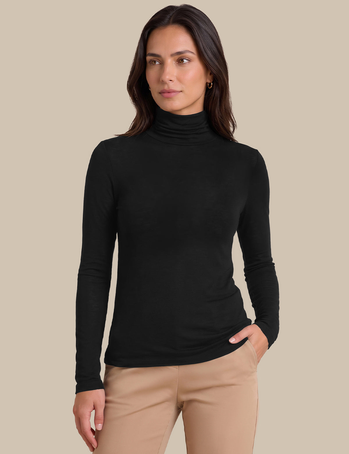 A woman modeling a black long-sleeve ribbed turtleneck top with beige pants, standing against a solid beige studio background.