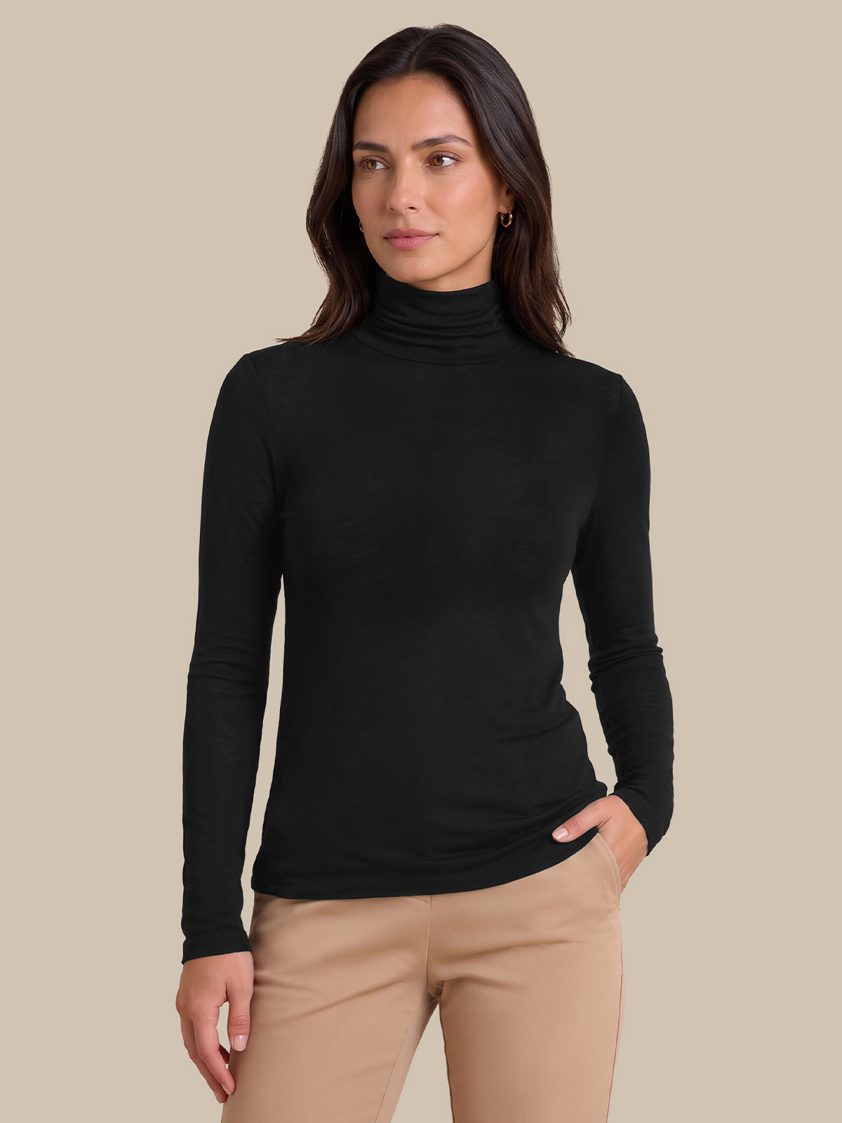 A woman modeling a black long-sleeve ribbed turtleneck top with beige pants, standing against a solid beige studio background.