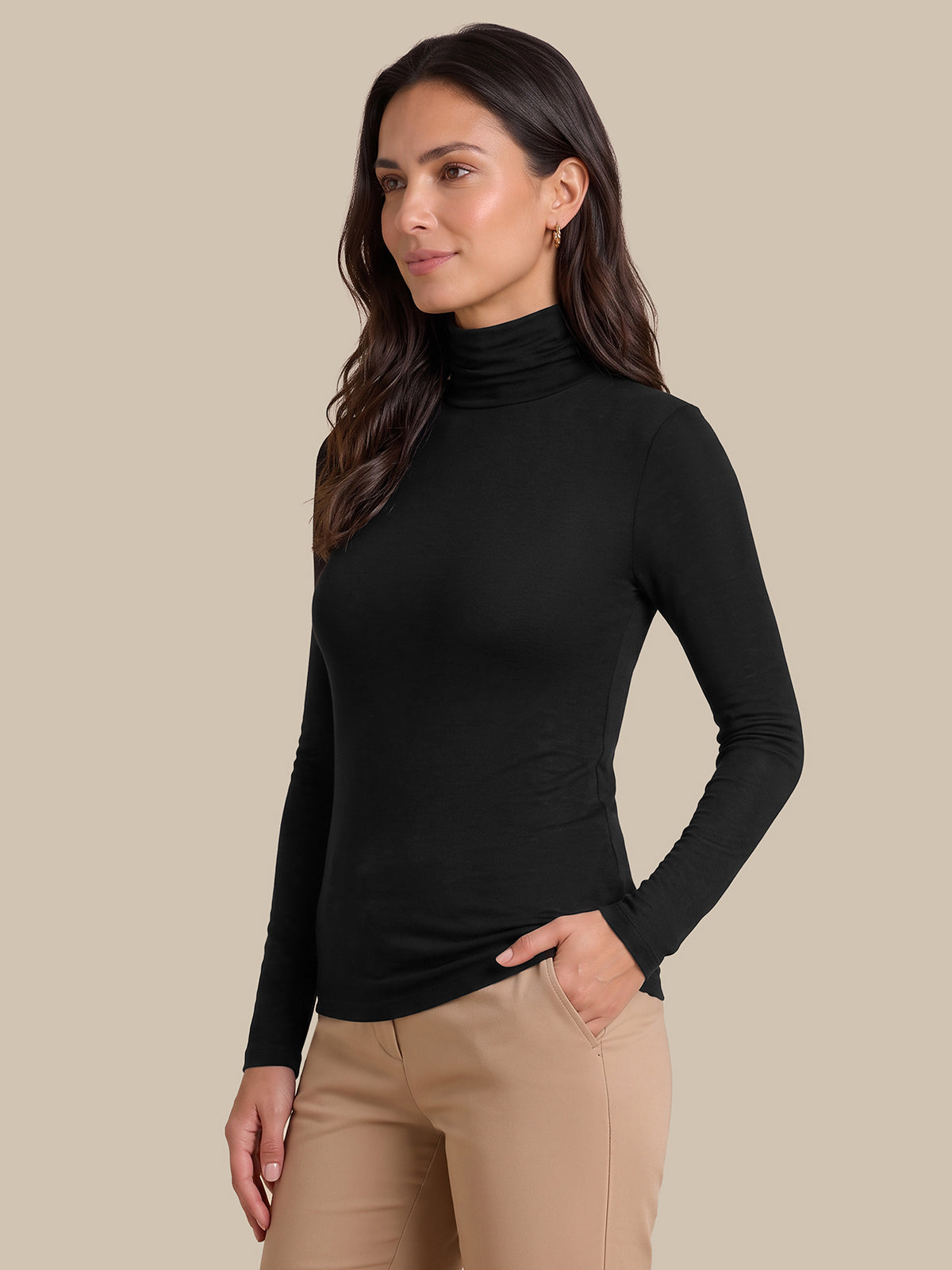 A woman modeling a fitted black long-sleeve turtleneck top with beige pants, standing against a solid beige studio background.