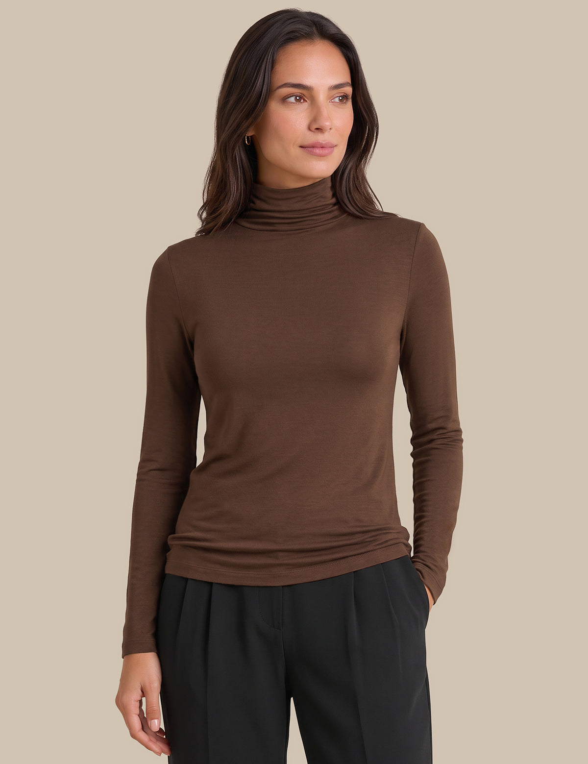 A woman wearing a fitted brown long-sleeve turtleneck top and black pants, standing against a plain beige studio background.