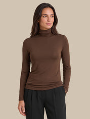 A woman wearing a fitted brown long-sleeve turtleneck top and black pants, standing against a plain beige studio background.