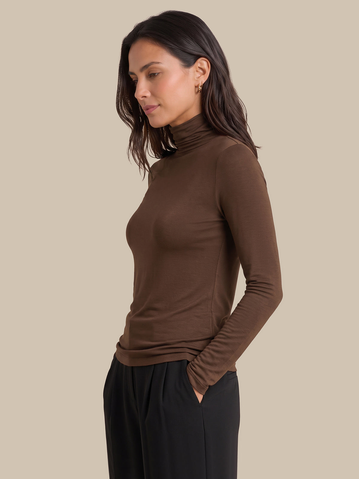 A woman wearing a fitted long-sleeve turtleneck top in dark brown, paired with black pants, shown in profile against a plain beige studio background.