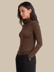 A woman wearing a fitted long-sleeve turtleneck top in dark brown, paired with black pants, shown in profile against a plain beige studio background.