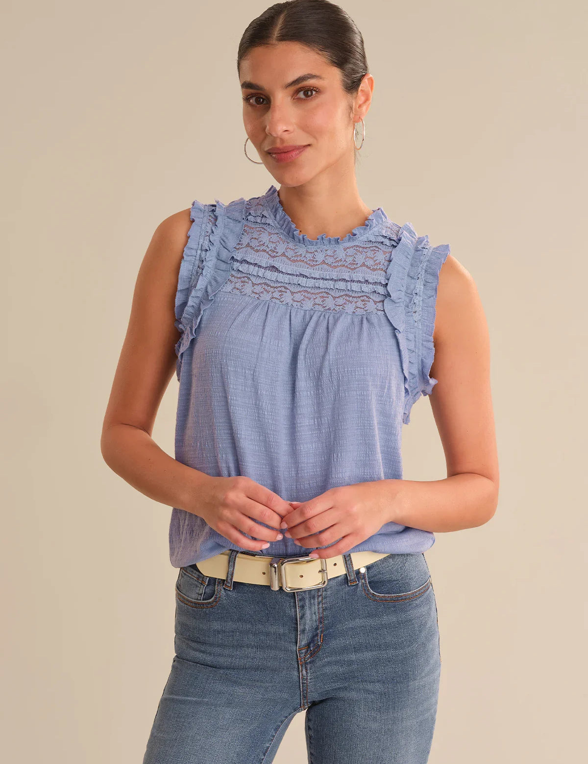 Woman wearing a blue sleeveless top with ruffled details and jeans on a beige background