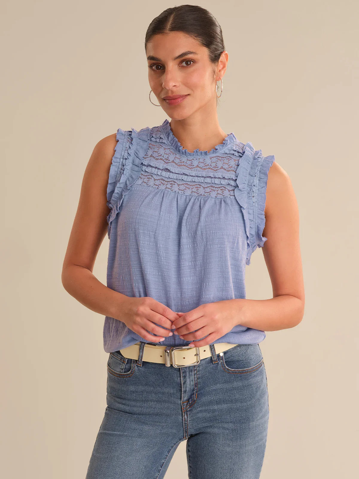 Woman wearing a blue sleeveless top with ruffled details and jeans on a beige background