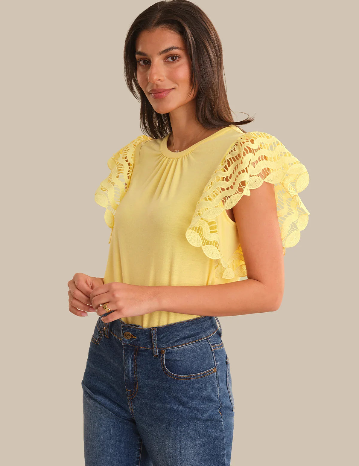 Woman wearing a yellow blouse with lace details and blue jeans on a beige background