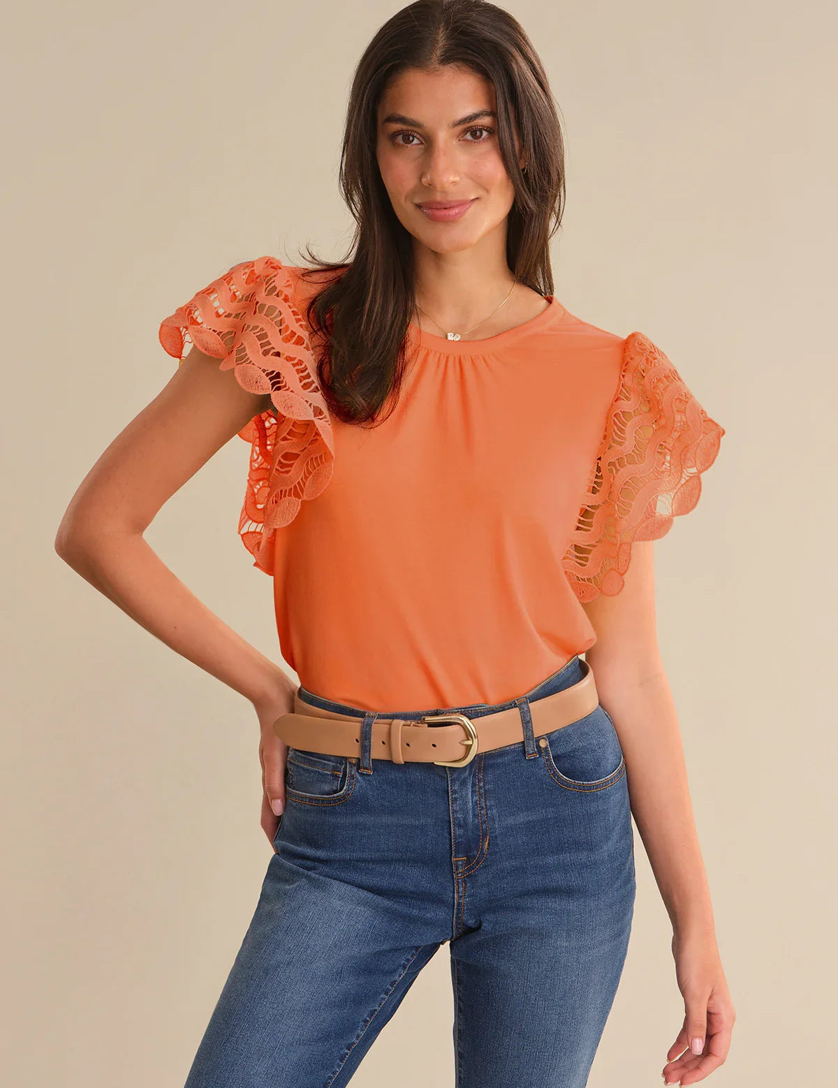 Woman wearing an orange top with lace sleeves and blue jeans on a beige background
