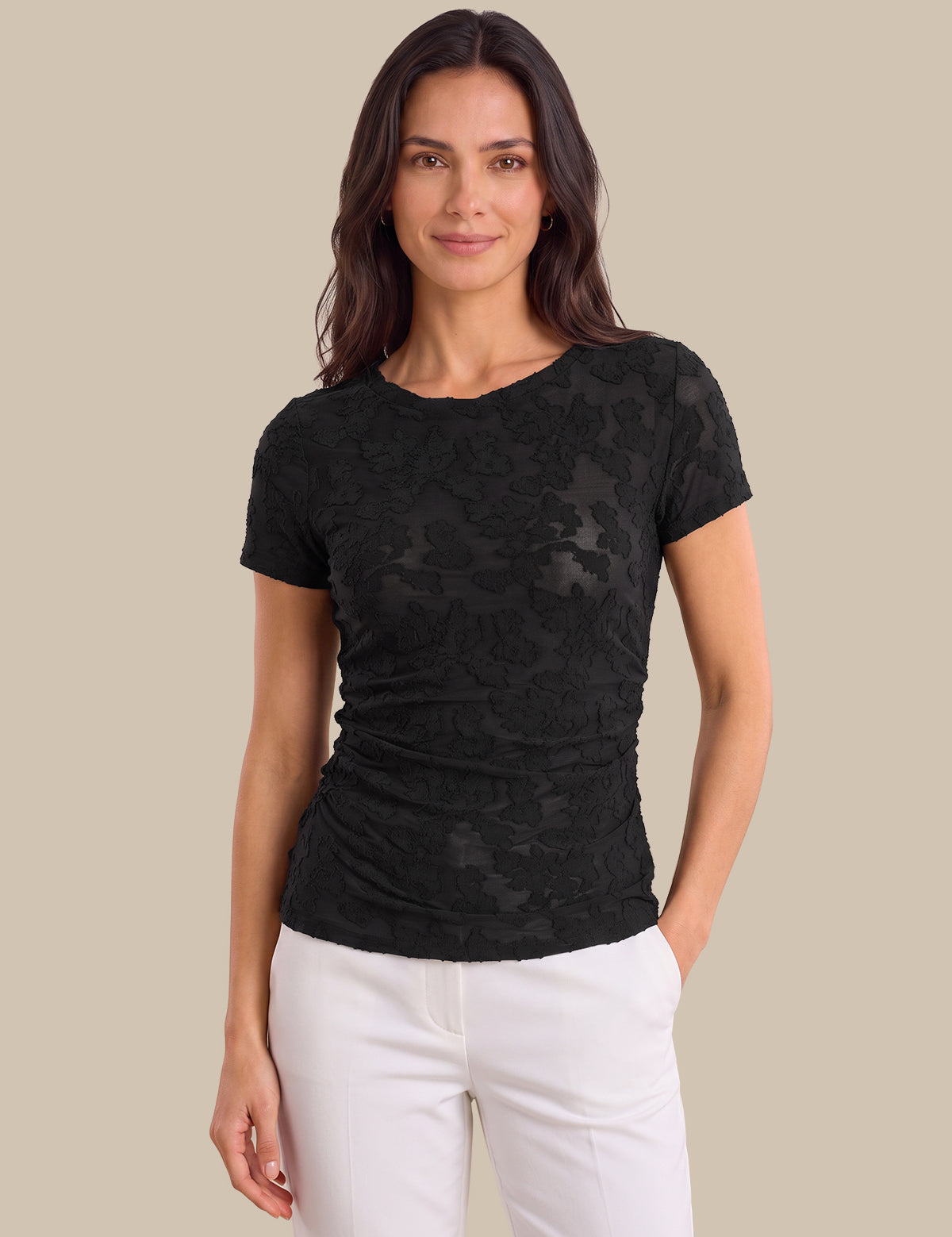 A woman modeling a black short-sleeve t-shirt with a textured, slightly sheer floral lace-like pattern, paired with white pants, against a solid beige background.