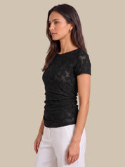 A woman wearing a black short-sleeve lace top and white pants, shown in profile against a solid beige background.