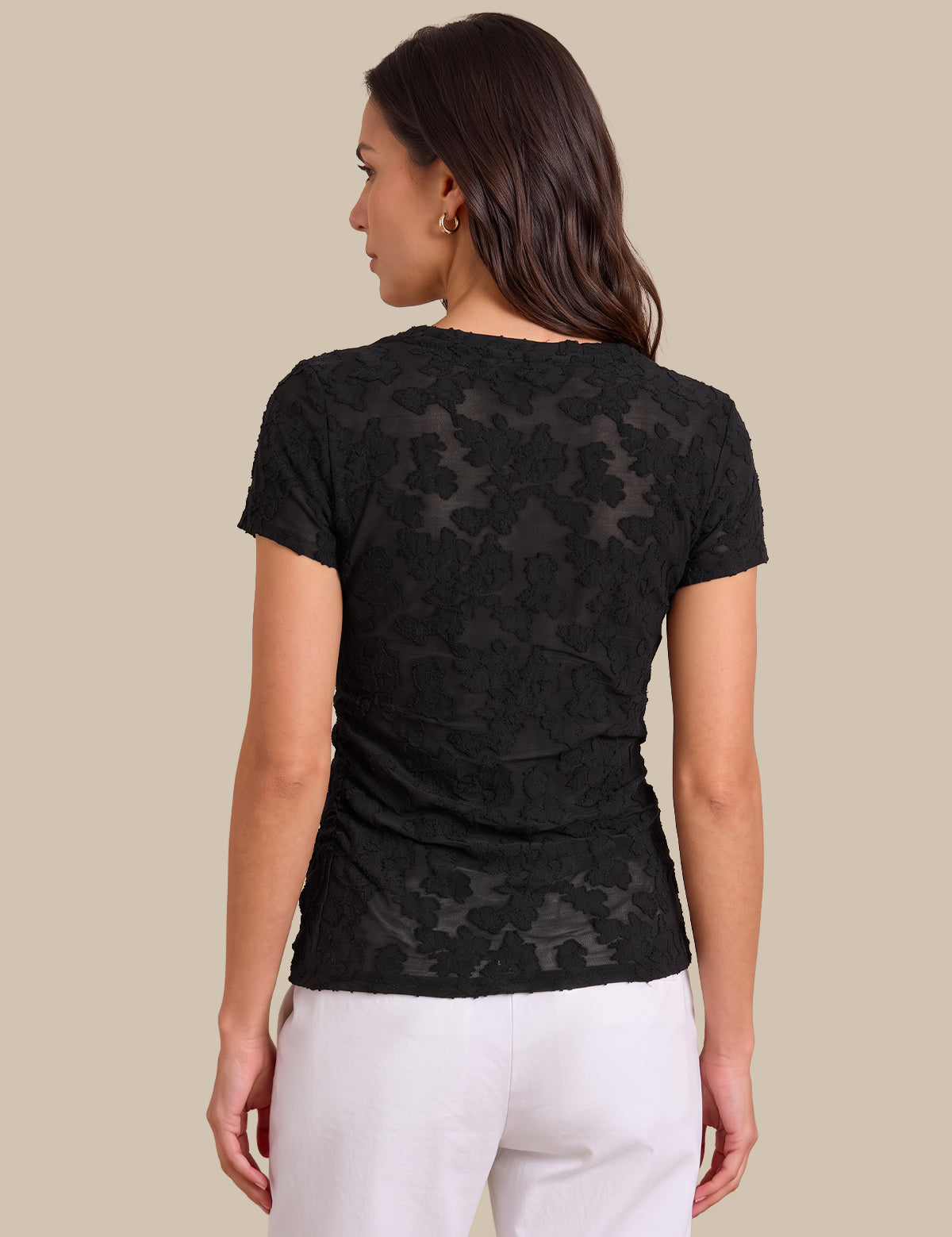 A woman shown from the back wearing a black short-sleeve lace top and white pants, photographed against a plain beige studio background.