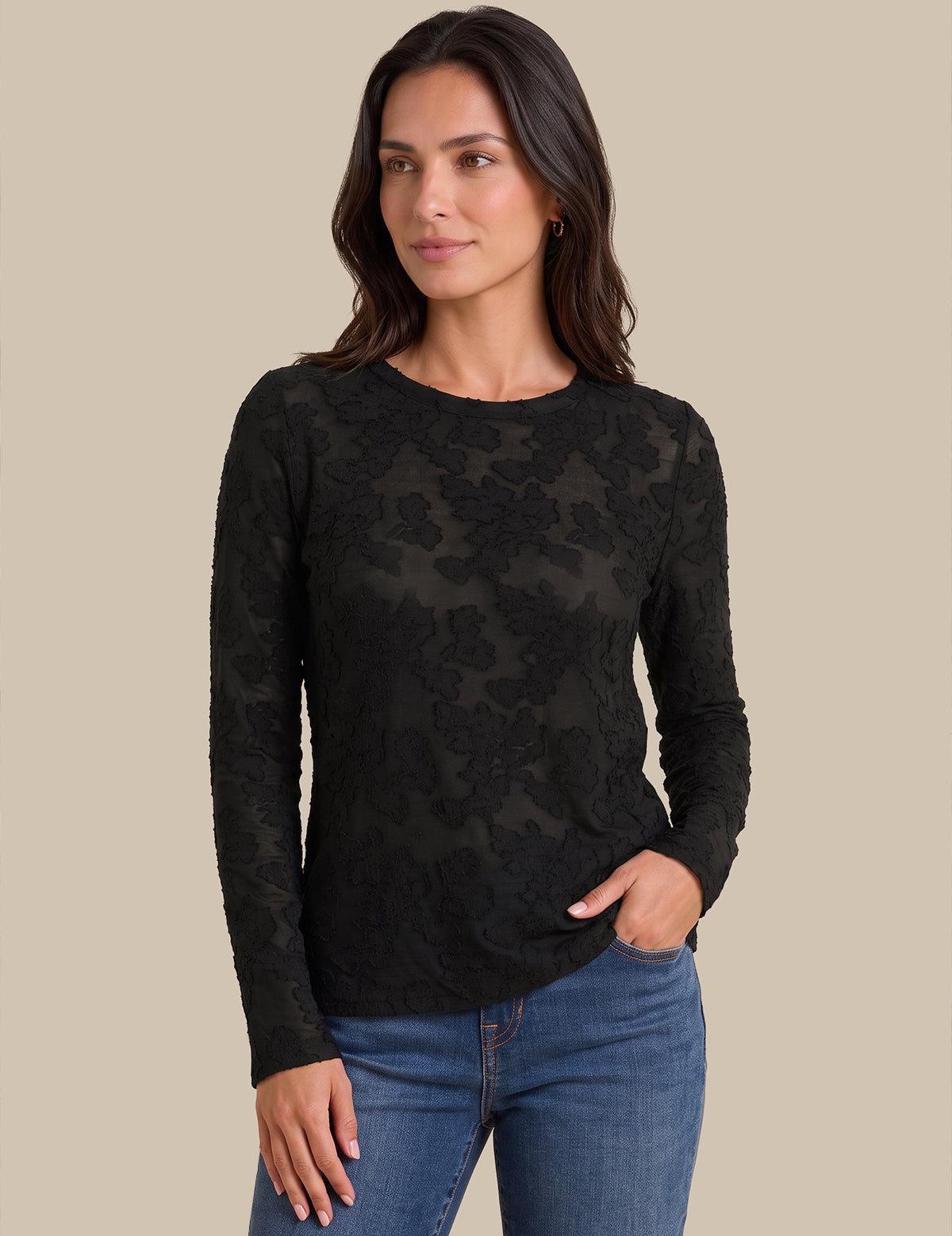 A woman modeling a black long-sleeve crewneck top with a sheer floral lace overlay, paired with blue jeans, against a solid beige background.