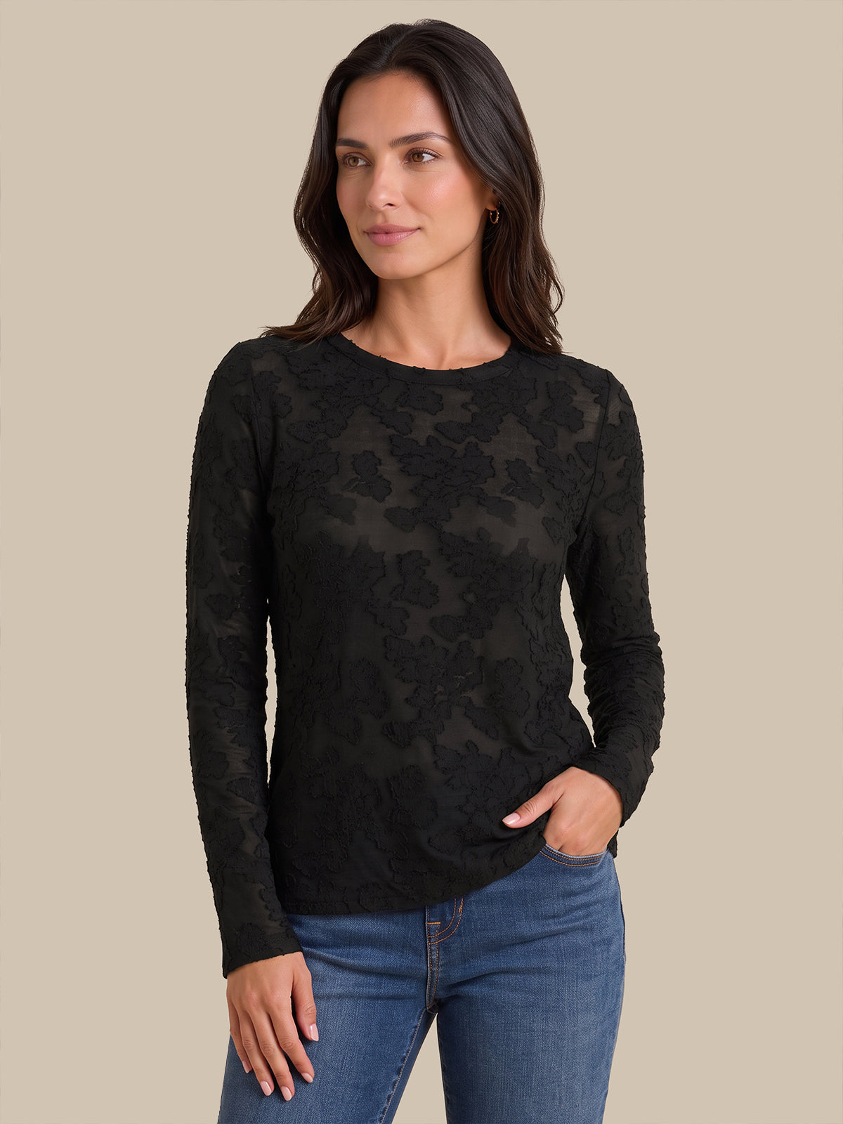 A woman modeling a black long-sleeve crewneck top with a sheer floral lace overlay, paired with blue jeans, against a solid beige background.