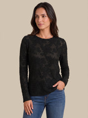 A woman modeling a black long-sleeve crewneck top with a sheer floral lace overlay, paired with blue jeans, against a solid beige background.