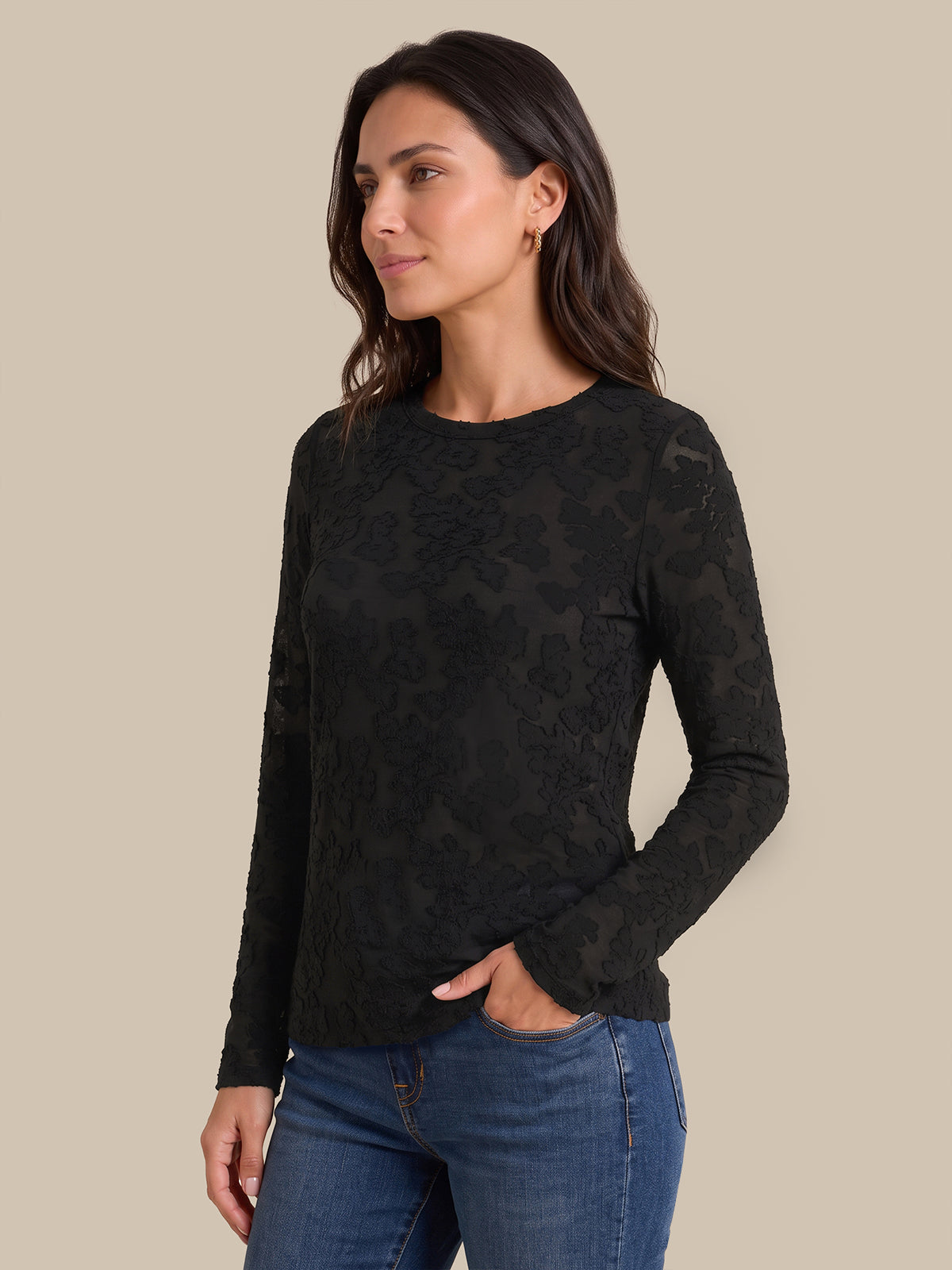 A woman wearing a black long-sleeve crewneck sweater with a subtle tonal floral lace pattern, paired with blue jeans, set against a solid beige background.