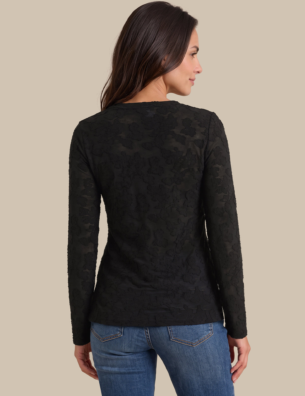 A woman shown from the back wearing a black long-sleeve knit top with an allover tonal floral lace pattern, paired with blue jeans, against a solid beige background.