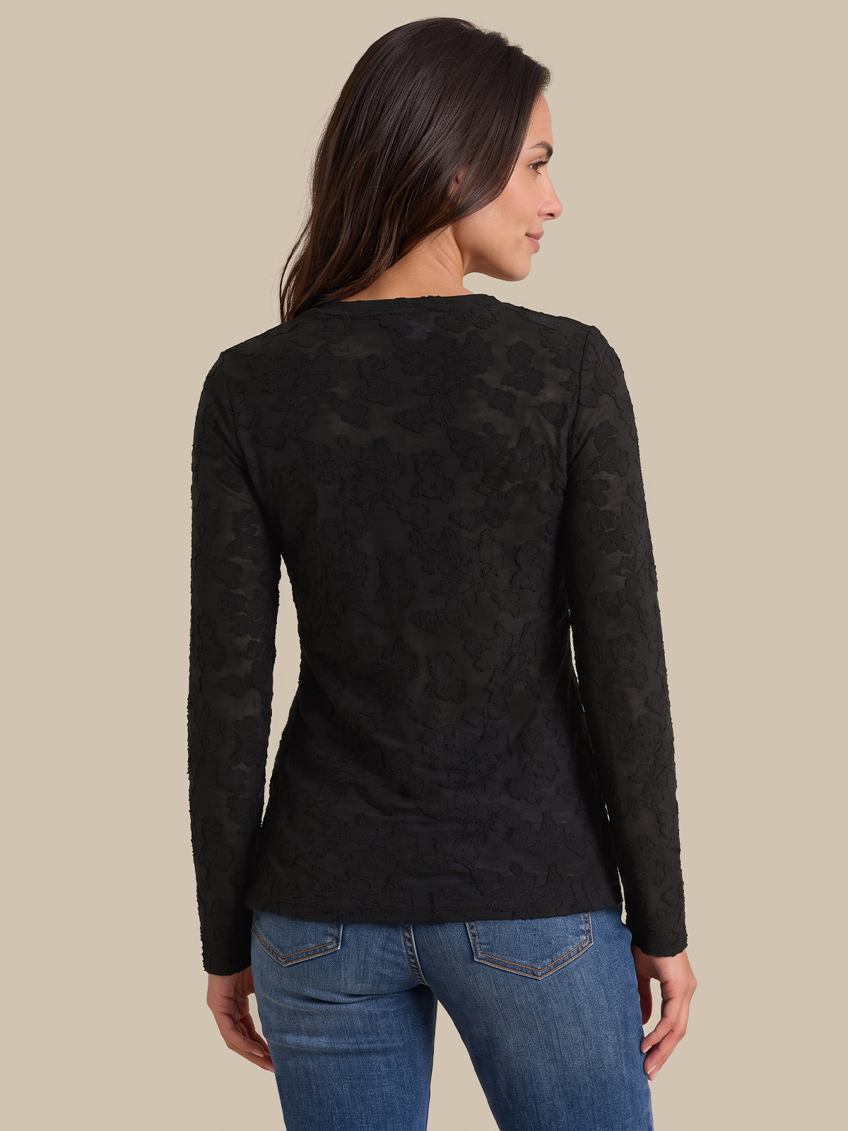 A woman shown from the back wearing a black long-sleeve knit top with an allover tonal floral lace pattern, paired with blue jeans, against a solid beige background.