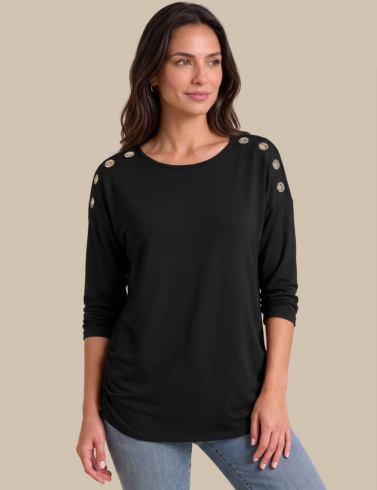 A woman modeling a black long-sleeve top with decorative gold-toned buttons on the shoulders, paired with light blue jeans, against a solid beige background.