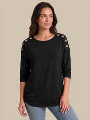 A woman modeling a black long-sleeve top with decorative gold-toned buttons on the shoulders, paired with light blue jeans, against a solid beige background.