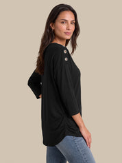 A woman modeling a black three-quarter sleeve top with decorative buttons on the shoulder, paired with blue jeans, against a solid beige background.