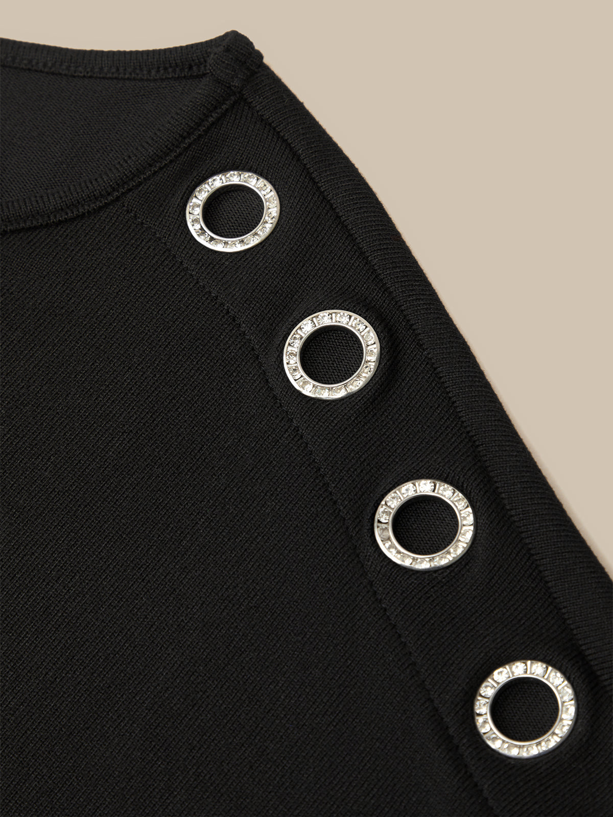 Close-up of a black knit garment featuring a vertical row of silver-tone metal grommets with clear rhinestone borders, set against a solid beige background.