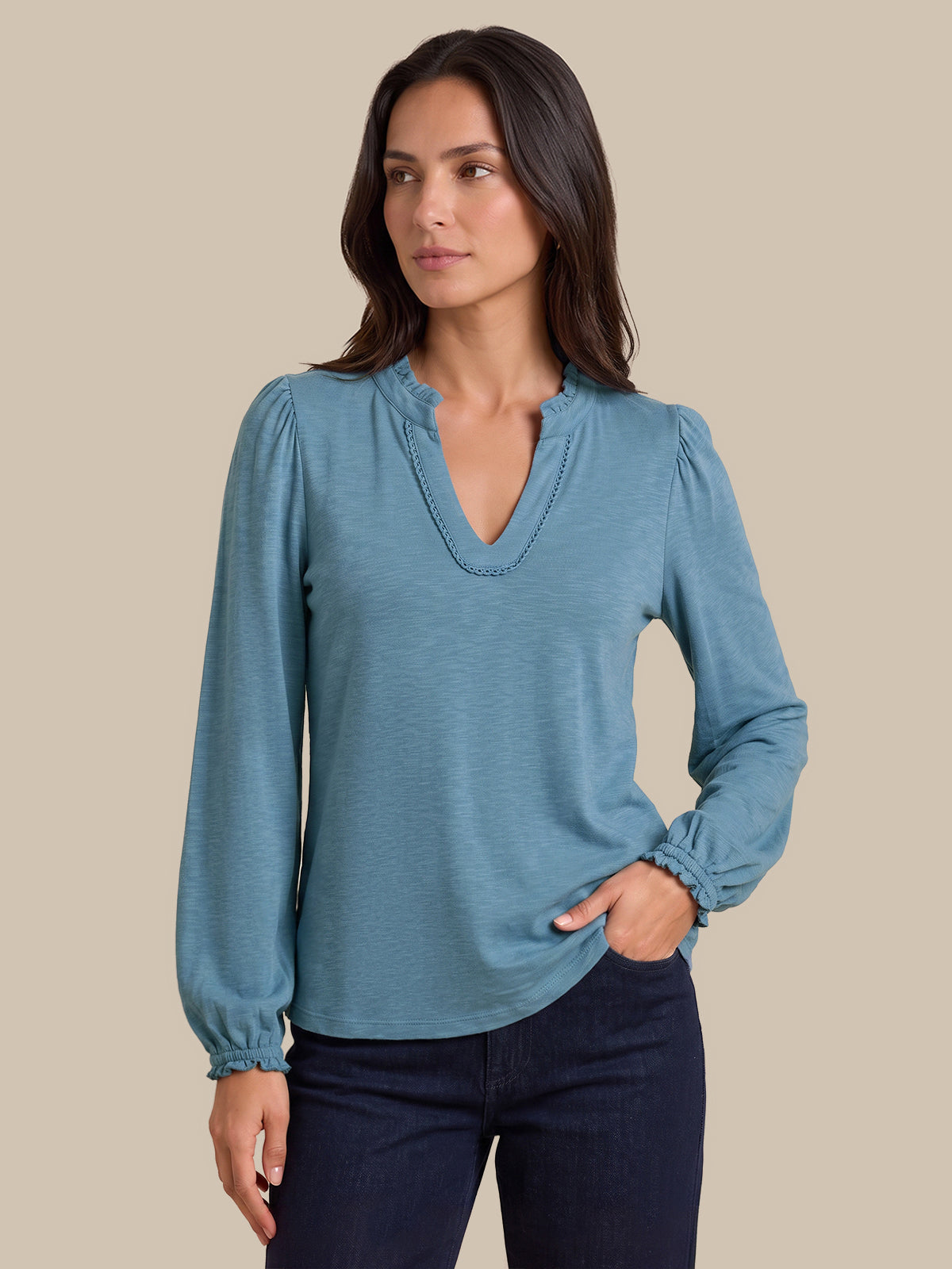 A woman modeling a dusty blue long-sleeve blouse with a split V-neckline and gathered cuffs, paired with dark blue jeans, against a solid beige background.