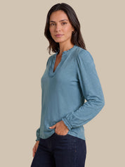 A woman modeling a dusty blue long-sleeve top with a ruffled split neckline and gathered cuffs, paired with dark blue jeans, against a solid beige background.