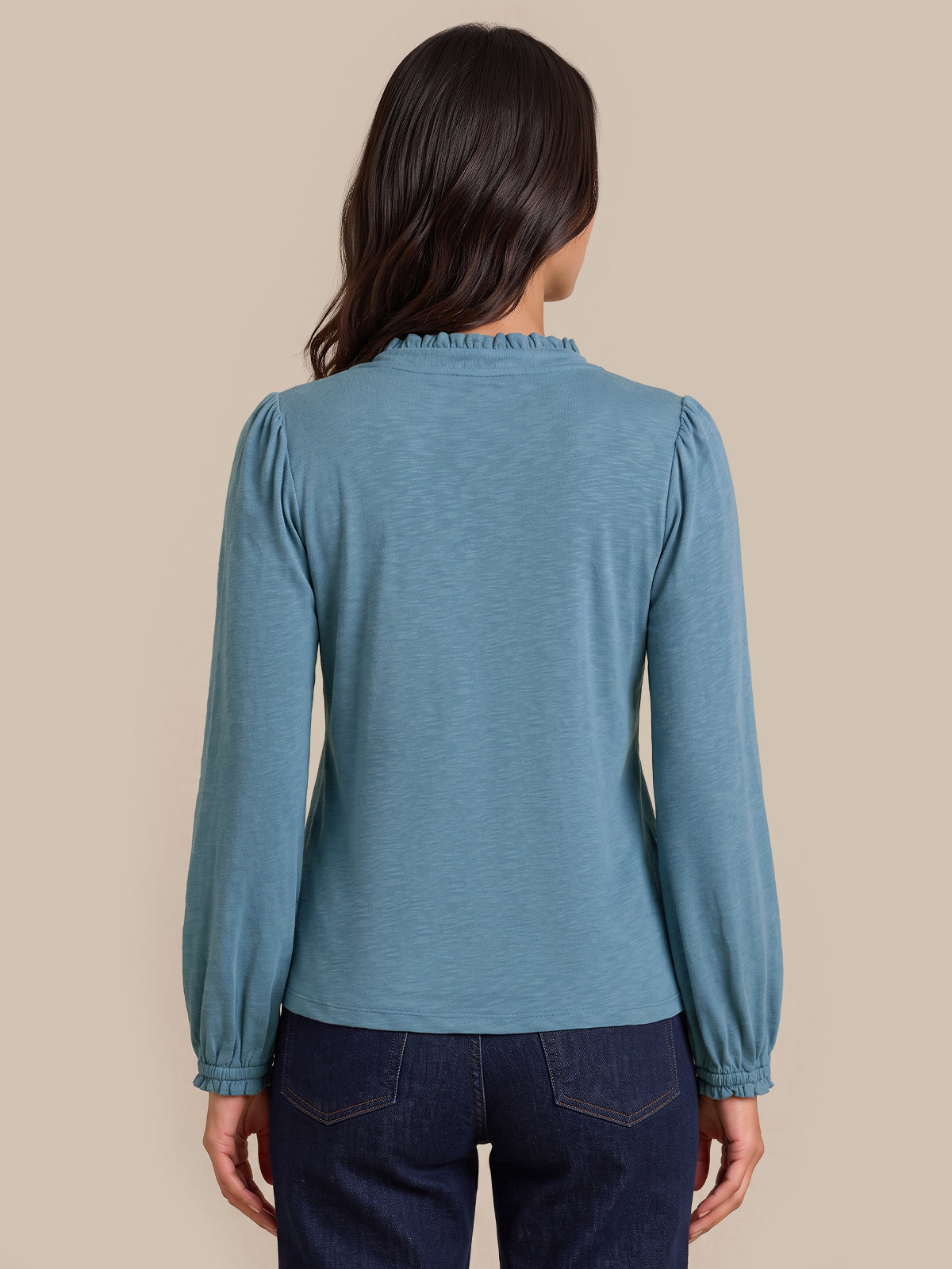 A woman viewed from the back wearing a slate blue long-sleeve top with a ruffled mock neck and gathered sleeves, paired with dark blue jeans, against a solid beige background.