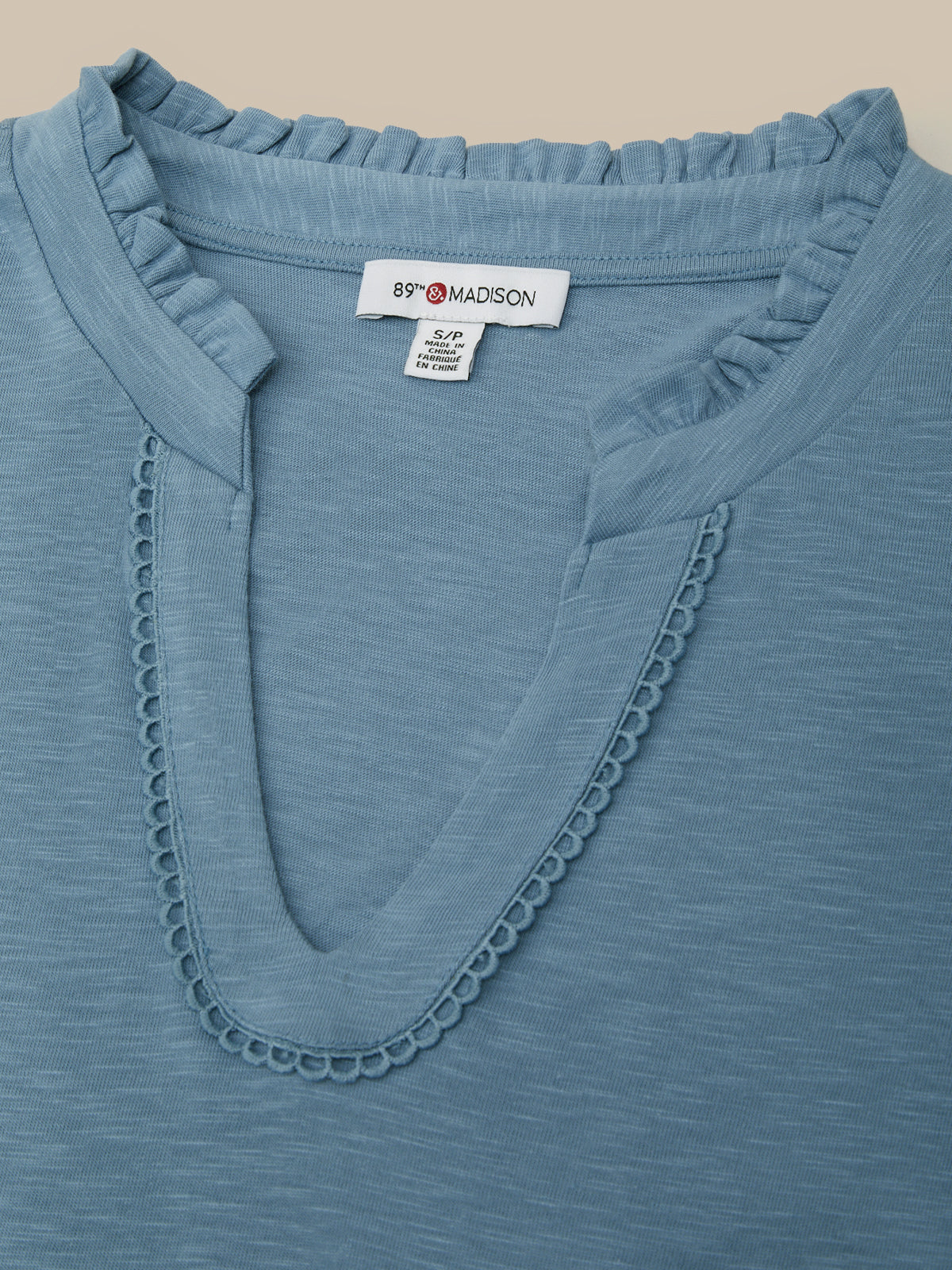 Close-up of a slate blue V-neck top with ruffled trim and scalloped neckline detailing, showing the brand label 'B'F*RD MADSON'.