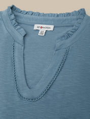 Close-up of a slate blue V-neck top with ruffled trim and scalloped neckline detailing, showing the brand label 'B'F*RD MADSON'.
