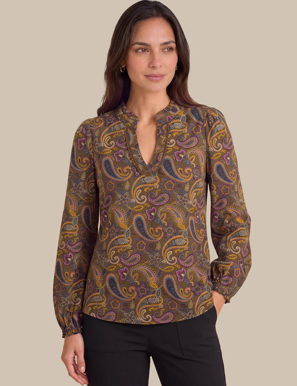 A woman modeling a long-sleeve blouse with a brown, gold, and purple paisley pattern and a split neckline, paired with black pants against a solid beige background.