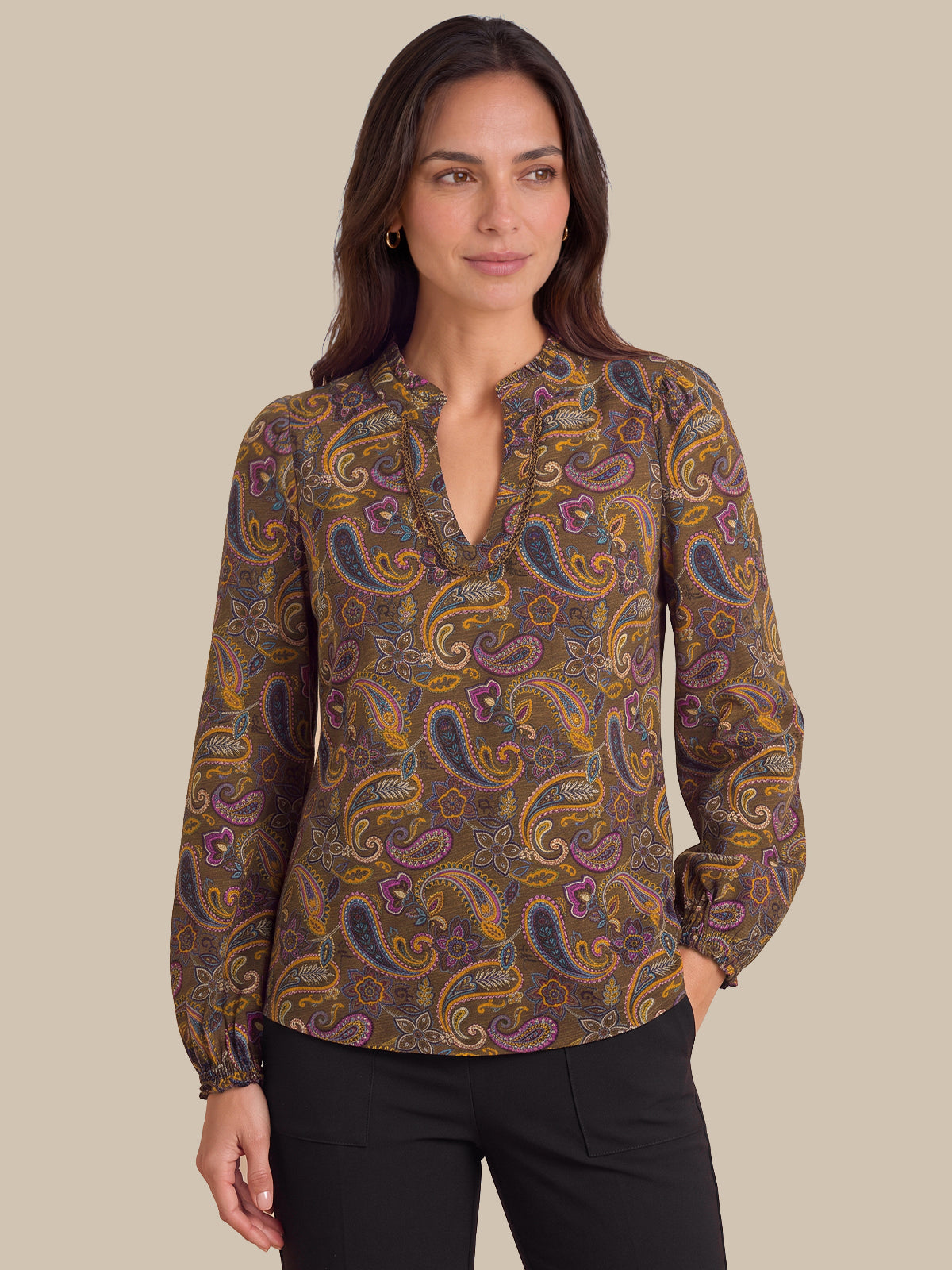 A woman modeling a long-sleeve blouse with a brown, gold, and purple paisley pattern and a split neckline, paired with black pants against a solid beige background.