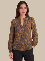 A woman modeling a long-sleeve blouse with a brown, gold, and purple paisley pattern and a split neckline, paired with black pants against a solid beige background.