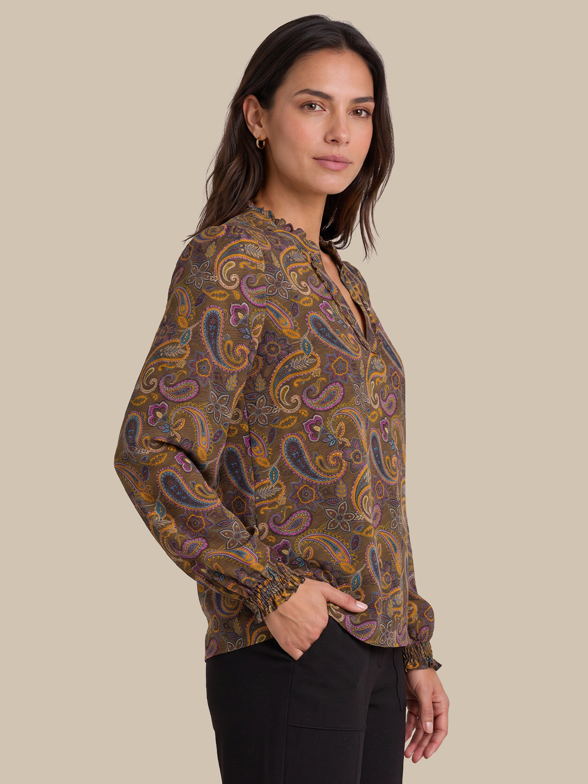 A woman wearing a long-sleeve paisley blouse with a brown, gold, and purple pattern, paired with black pants, photographed against a solid beige background.