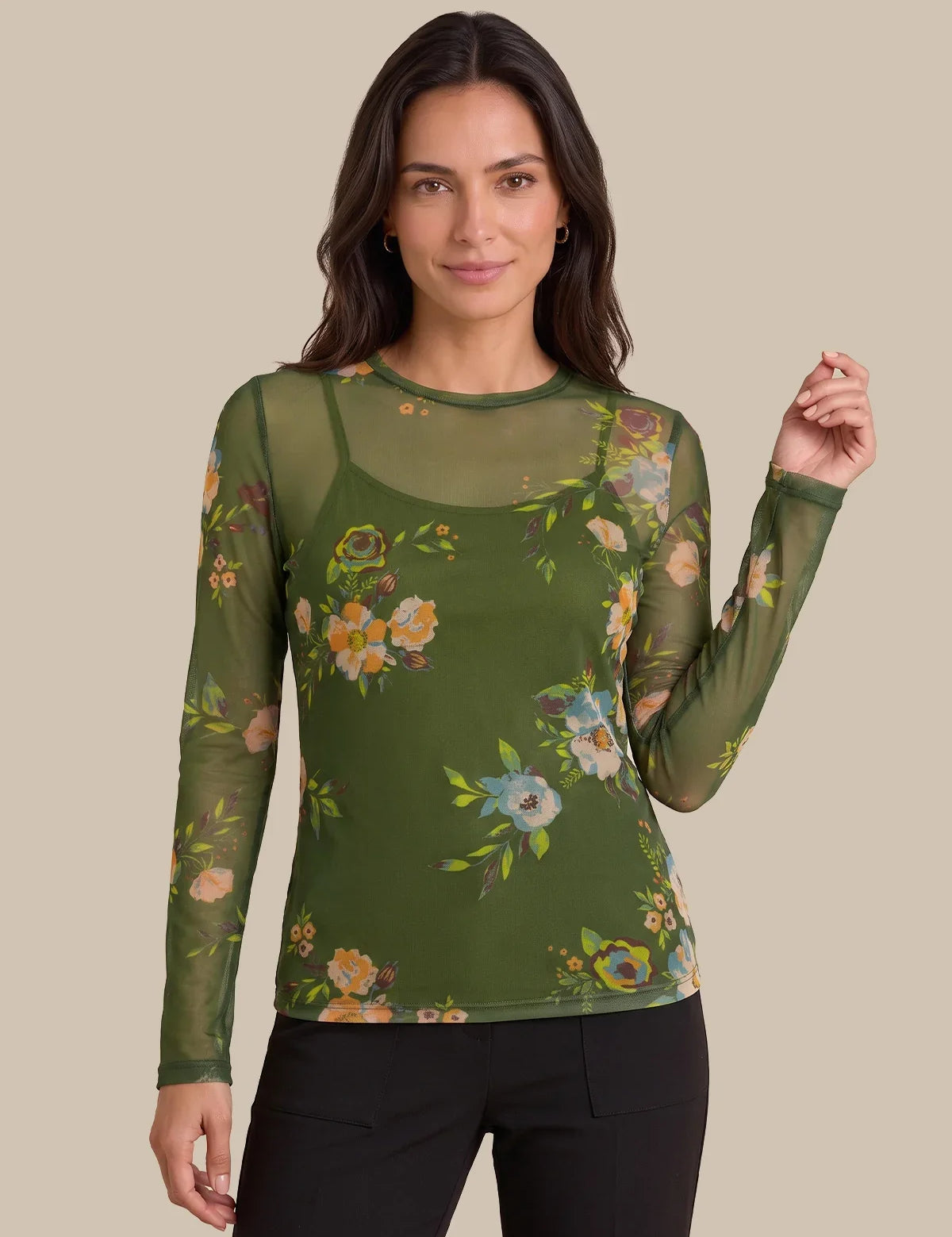 A woman modeling a sage green long-sleeve mesh top with a colorful floral print over a matching green camisole, paired with black pants, against a solid beige background.