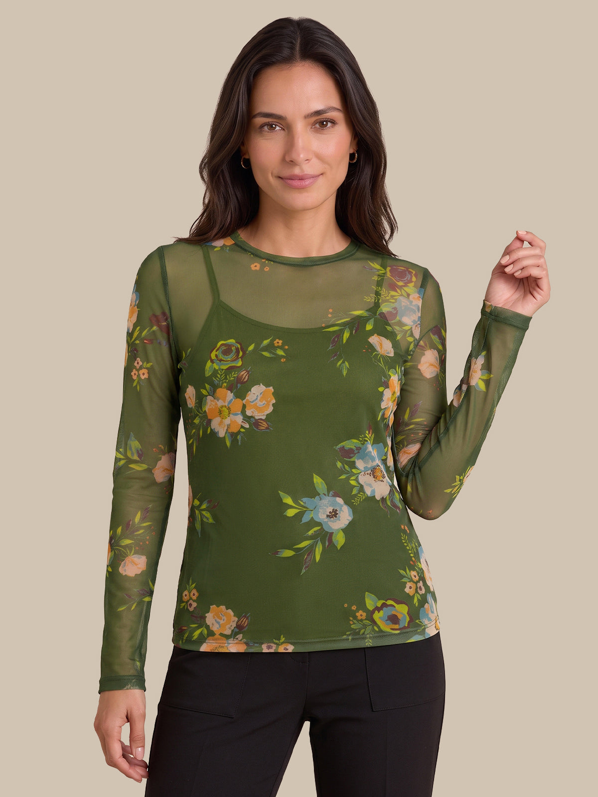 A woman modeling a sage green long-sleeve mesh top with a colorful floral print over a matching green camisole, paired with black pants, against a solid beige background.