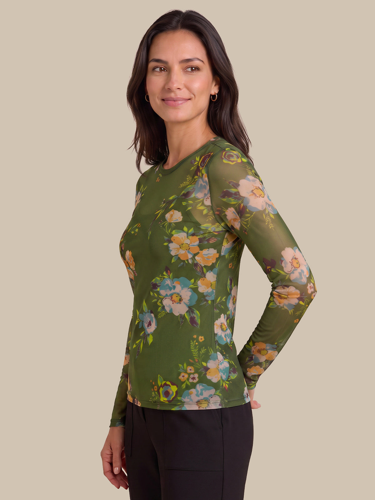 A woman modeling a long-sleeve sheer mesh top with an olive green base and a colorful floral print, paired with black pants, against a solid beige background.