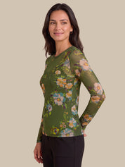 A woman modeling a long-sleeve sheer mesh top with an olive green base and a colorful floral print, paired with black pants, against a solid beige background.