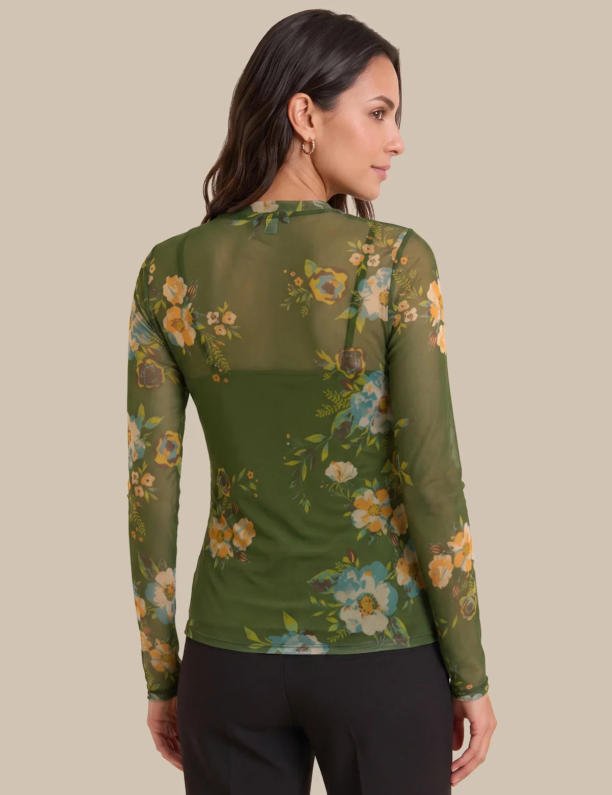 A woman shown from the back wearing a sheer olive green long-sleeve top with a floral print in yellow, blue, and white, paired with black pants, against a solid beige background.