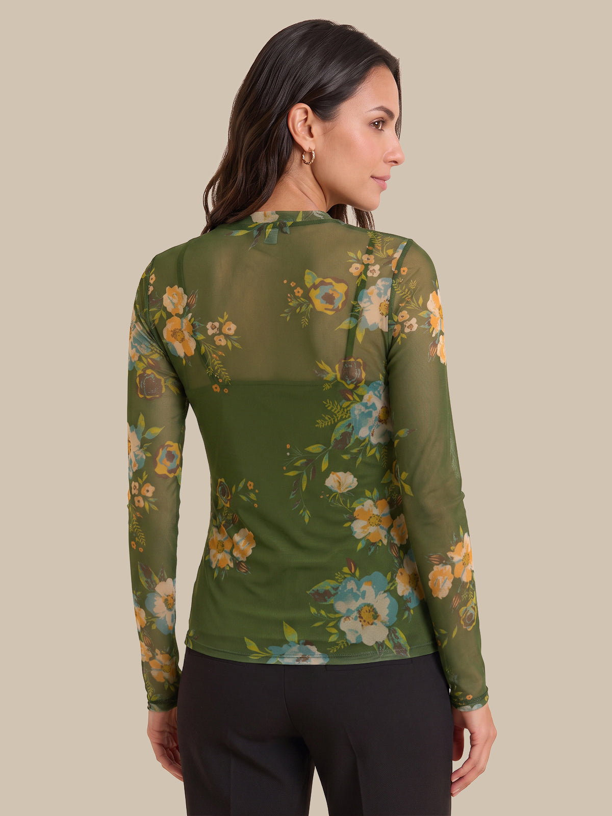A woman shown from the back wearing a sheer olive green long-sleeve top with a floral print in yellow, blue, and white, paired with black pants, against a solid beige background.